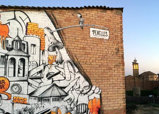 Un grafiti en una de las primeras casas del pueblo de Penelles con el campanario de la iglesia en el fondo.